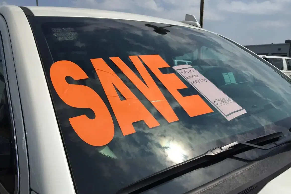 A car at a dealership with a large orange 'SAVE' sign on the windshield, promoting a special offer.
