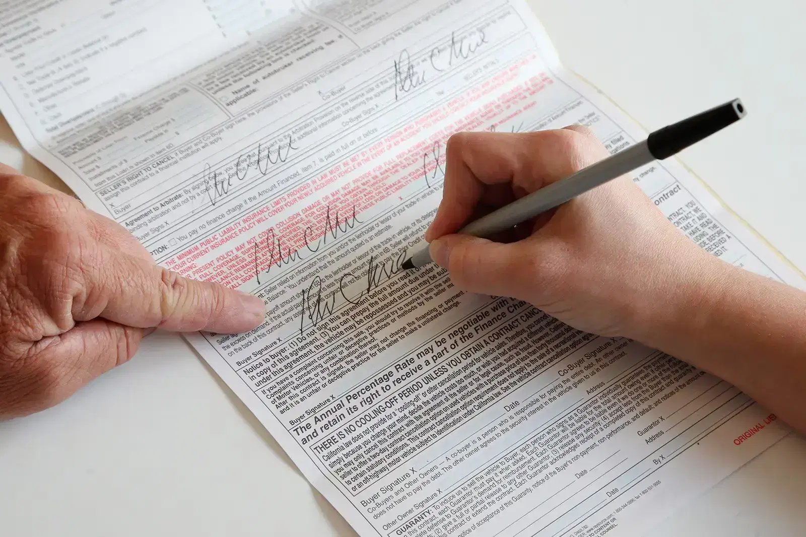 A close-up of a person signing a car purchase contract while a dealer points to important terms on the document.