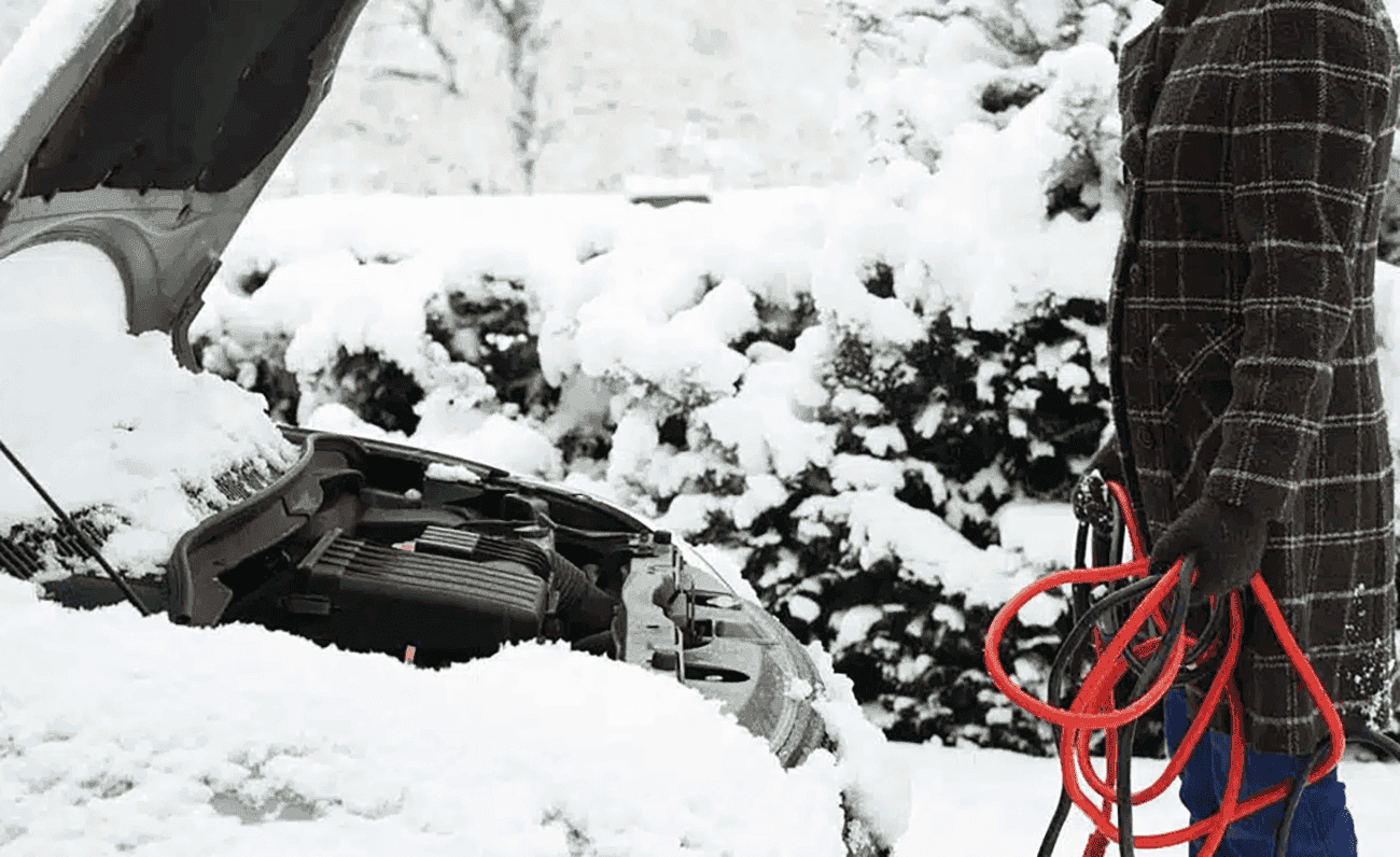 A person holding jumper cables next to a car with an open hood, struggling with a dead battery in snowy weather.