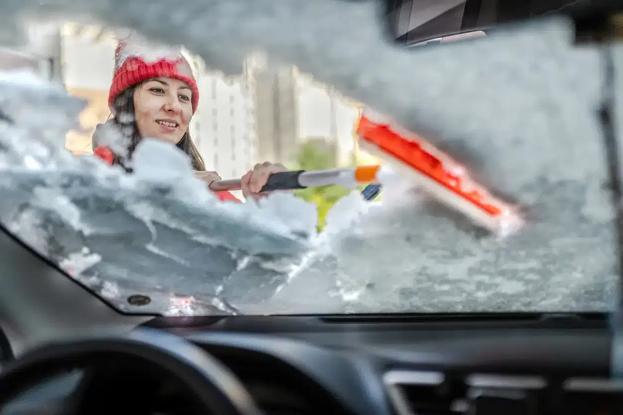 Woman in winter jacket scraping ice and snow from her car windows on a freezing morning.